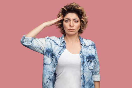 Yes sir. Portrait of serious young worker woman with curly hairstyle in casual blue shirt standing and looking at camera with salute and ready to do smoething. studio shot, isolated on pink background