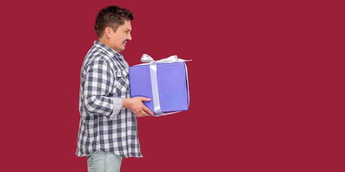 Side view portrait of happy modern middle aged man in white t-shirt and in checkered shirt standing and giving you big heavy gift box with toothy smile. indoor studio shot on isolated red background.