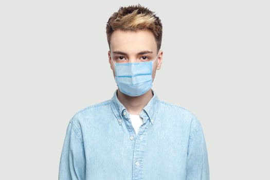 Portrait of calm serious handsome young man with surgical medical mask in light blue shirt standing and looking at camera with serious face. indoor studio shot on grey background copy space.
