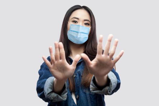 Portrait of scared beautiful asian young woman with surgical medical mask in blue denim jacket standing with hand blocking and looking at camera. indoor studio shot, isolated on grey background