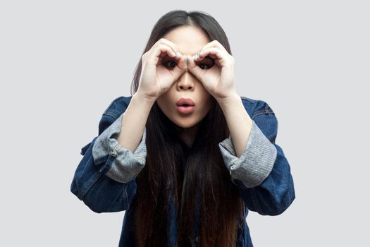 Portrait of amazed beautiful brunette asian girl in casual blue denim jacket with makeup standing with binoculars hand gesture on eyes and looking. studio shot, isolated on light grey background.