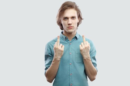 Portrait of poker face handsome long haired blonde young man in blue casual shirt standing and looking at camera with middle finger fuck sign. indoor studio shot, isolated on light grey background.