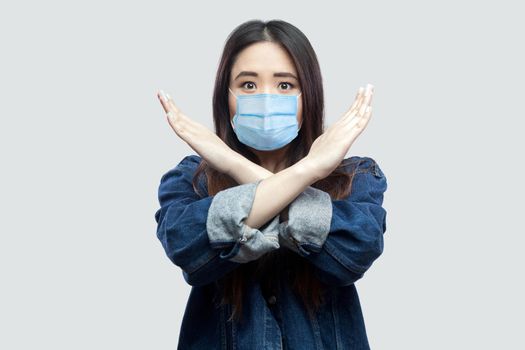 Portrait of worried brunette asian young woman with surgical medical mask in casual blue denim jacket standing and showing closed or x sign at camera. indoor studio shot, isolated on grey background.