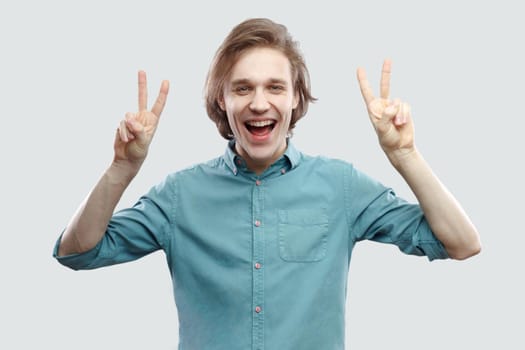 Portrait of happy handsome long haired blonde young man in blue shirt standing and looking at camera with amazing face, victory or peace gesture. indoor studio shot, isolated on light grey background.