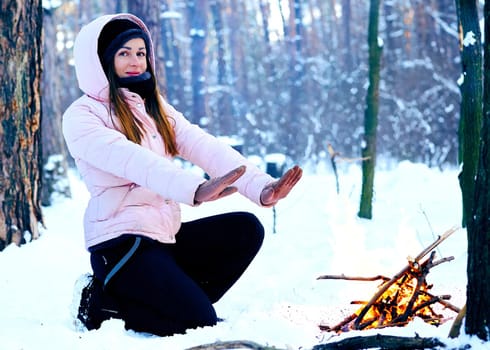 a large open-air fire used as part of a celebration, for burning trash, or as a signal. Young woman on a winter picnic in the forest warming up near the fire.