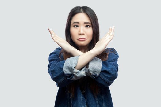 Portrait of worried beautiful brunette asian young woman in casual blue denim jacket with makeup standing and showing closed or x sign at camera. indoor studio shot, isolated on light grey background.