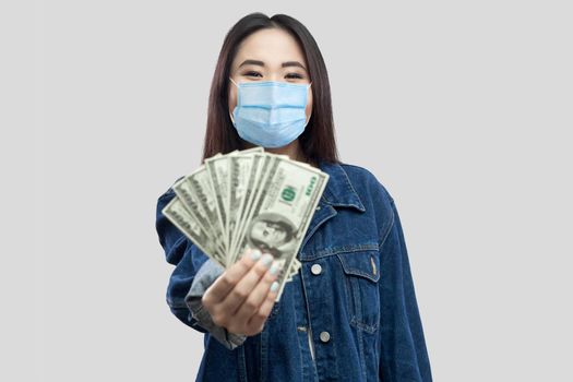 Portrait of successful attractive young asian woman with medical mask standing, holding dollars, demonstration and looking at camera with smile. Indoor, studio shot, isolated, gray background