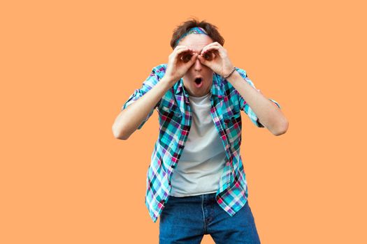 Portrait of surprised young man in casual blue checkered shirt and headband standing with binoculars hands gesture on eyes, looking with shocked face. indoor studio shot, isolated on orange background