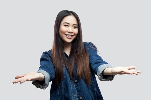 Portrait of happy beautiful brunette asian young woman in blue jacket with makeup standing looking at camera with raised arms to share and toothy smile. studio shot, isolated on light grey background.