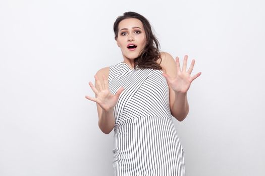 Portrait of scared beautiful young brunette woman with makeup and striped dress standing and looking at camera with blocking hands. indoor studio shot, isolated on grey background.