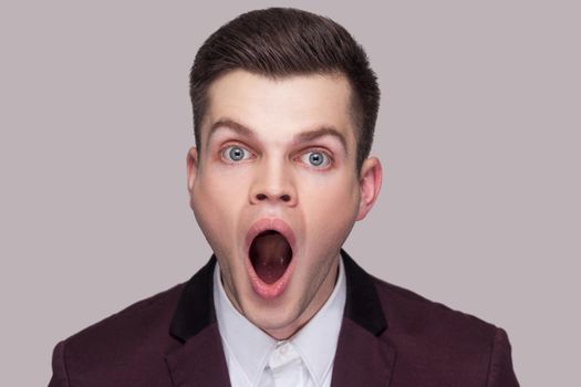 Closeup portrait of shocked handsome young man in violet suit and white shirt, standing, looking at camera with open mouth and shouting. indoor studio shot, isolated on grey background.