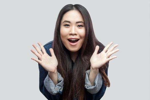 Portrait of surprised beautiful brunette asian young woman in blue jacket with makeup standing with raised arms and looking at camera with excited face. studio shot, isolated on light grey background.