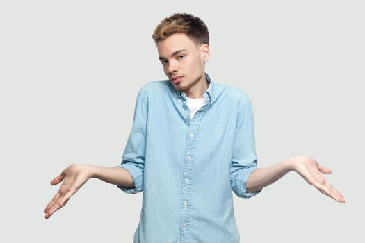 I don't know. Portrait of confused handsome young man in light blue shirt standing with puzzled face, raised arms and looking at camera. indoor studio shot on grey background copy space.