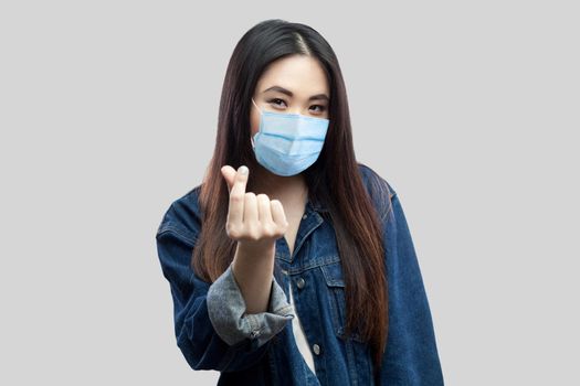Portrait of funny brunette asian young woman with medical mask in casual blue denim jacket standing and showing money gesture and looking at camera. studio shot, isolated on grey background.