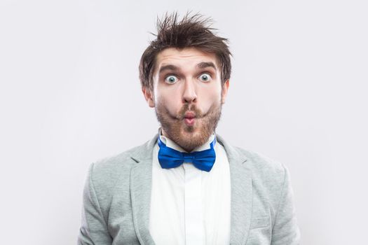 Closeup portrait of funny surprised handsome bearded man in casual grey suit, blue bow tie standing and looking at camera with big eyes and fish lips. studio shot, isolated on light grey background.