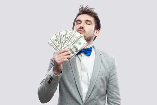 Portrait of satisfied attractive young businessman in gray coat and blue bow tie standing, kissing with closed eyes and demonstrating fan of money. Indoor studio shot, isolated, gray background.