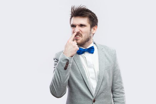 Your are liar. Portrait of angry handsome bearded man in casual grey suit and blue bow tie standing and touching his nose with lie gesture. indoor studio shot, isolated on light grey background.