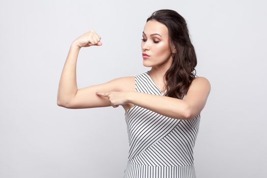 I am strong. Portrait of proud beautiful young brunette woman with makeup and striped dress standing and pointing at her biceps. indoor studio shot, isolated on grey background.