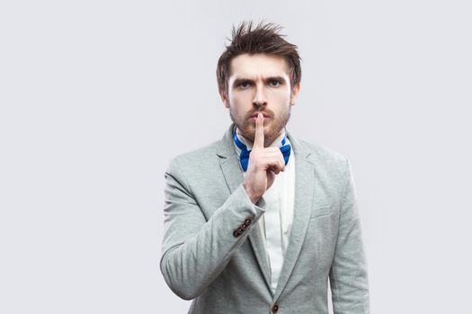 Portrait of serious handsome bearded man in casual grey suit and blue bow tie standing, looking at camera with silence sign. indoor studio shot, isolated on light grey background.