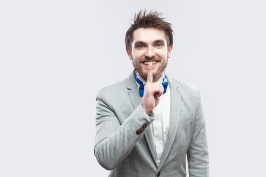 This is secret between us. Portrait of handsome bearded man in casual grey suit and blue bow tie standing with silent sign and toothy smile. indoor studio shot, isolated on light grey background.