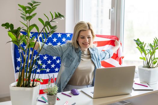 Happy woman employee sitting wrapped in USA flag, shouting for joy in office workplace, celebrating labor day or US Independence day. Indoor studio studio shot isolated on yellow background.