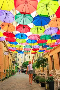 Street decoration. woman standing under colored umbrellas hanging at top. Set of different umbrellas. Local landmark. Cafe decoration in Lodz. Multicolored umbrella. Place of interest. Bold colors