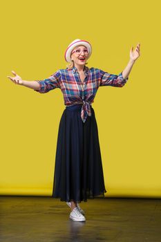 full length portrait of happy modern stylish mature woman in casual style with hat and eyeglasses standing with raised arms and surprised face looking. indoor studio shot on yellow wall background.