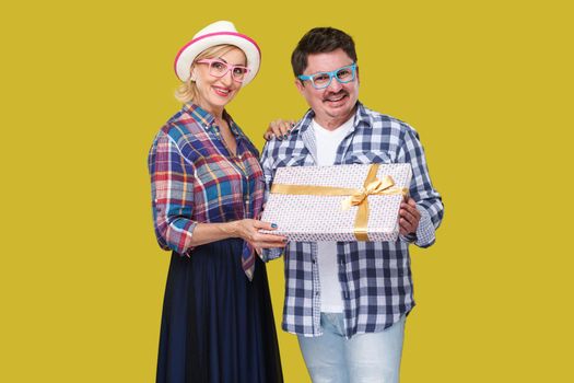 Couple of positive friends, adult man and woman in casual checkered shirt standing beside together holding present gift box, toothy smile and looking. Indoor, isolated, studio shot, yellow background