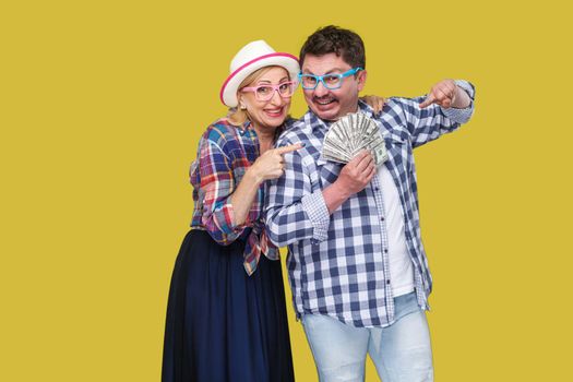 Happy wealthy family, adult man and woman in casual checkered shirt standing pickaback together, holding fan of dollar and pointing finger to money. Indoor,isolated, studio shot, yellow background