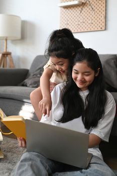 Cheerful young woman and her younger sister surfing internet with laptop computer at home.