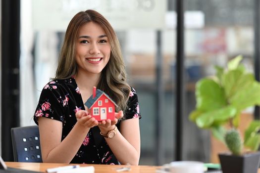 Happy young woman holding small house model and smiling at camera. Real estate investment concept.
