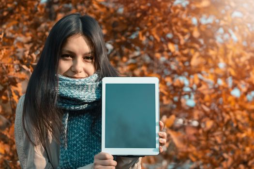 Girl shows the screen of the tablet to the viewer while standing against the backdrop of bright autumn foliage in the park.