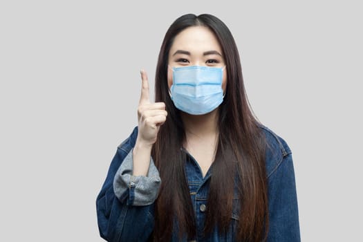 Portrait of exicted beautiful brunette asian girl with surgical medical mask in blue denim jacket standing and looking at camera , finger up. indoor studio shot, isolated on grey background.