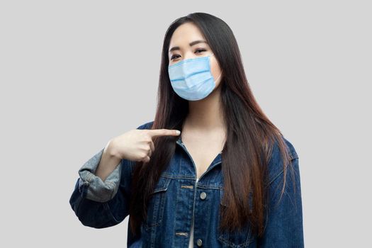 Portrait of proud satisfied beautiful brunette asian young woman with surgical medical mask in blue denim jacket standing pointing herself looking at camera. studio shot, isolated on grey background