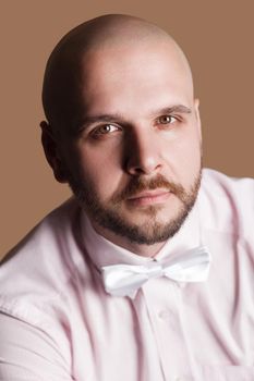 closeup portrait of handsome bearded bald man in light pink shirt and white bow, looking at camera with serious face. indoor studio shot, isolated on brown background.