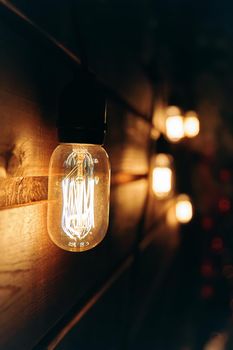 Vintage incandescent Edison type bulb on wooden texture wall. Christmas night party mood. Blurred warm garland light on background. Holiday decorating.