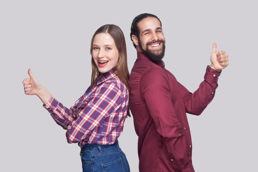 Profile side view portrait of happy satisfied bearded man and woman in casual style standing and looking at camera, smiling with thumbs up. indoor studio shot, isolated on gray background.