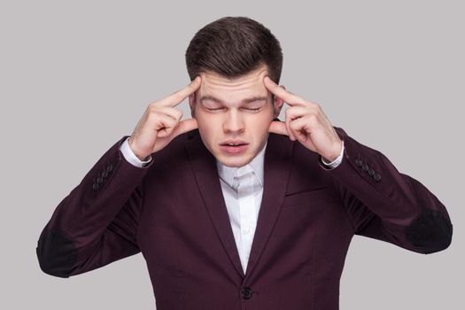 Portrait of upset handsome young man in violet suit and white shirt, standing with closed eyes and touching his painful head. indoor studio shot, isolated on grey background.