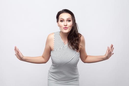 Portrait of happy lovely beautiful young brunette woman with makeup and striped dress standing with crossed arms and looking at camera with kiss. indoor studio shot, isolated on grey background.