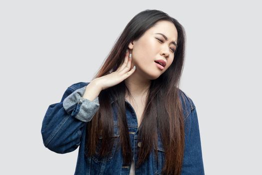 Neck pain. Portrait of sick beautiful brunette asian young woman in casual blue denim jacket standing feeling discomfort and touching her neck . indoor studio shot, isolated on light grey background.