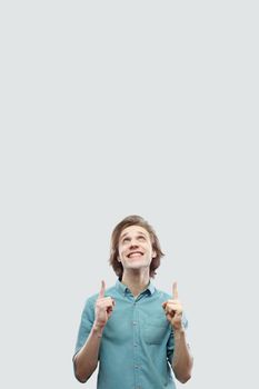 Portrait of happy toothy smiley handsome long haired blonde young man in blue casual shirt standing and pointing at up copyspace. indoor studio shot, isolated on light grey background.