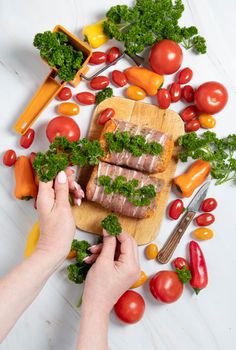 female hands decorate with parsley minced meat sausages in bacon with fresh vegetables, cherry tomatoes, sweet peppers, colorful food flat lay. High quality photo