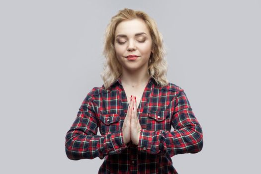 Yoga time. Portrait of calm beautiful blonde young woman in casual red checkered shirt standing with crossed arms and closed eyes, meditating. indoor studio shot, isolated on grey background.
