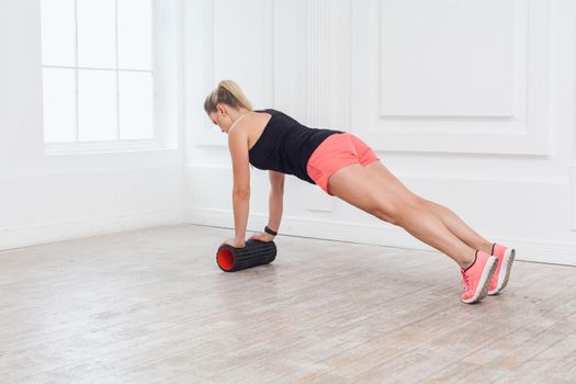 Back side view portrait of athletic blonde young woman in fit wear using foam roller in gym to workout to loss weight and doing plank. stretching and massaging muscles. Indoor, studio shot.
