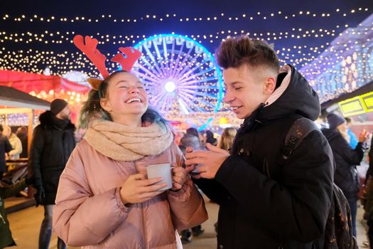 Christmas time, New Year holidays. Young people, couple of teenagers having fun at Christmas market, drinking hot tea from mugs, talking, laughing, background garlands of evening city, Ferris wheel