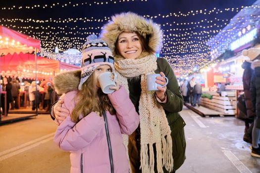 Christmas and New Year holidays, happy mom and daughter kid walking together drinking hot mug tea at Christmas market, sparkling lights of garlands of evening city ferris wheel