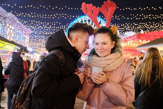 Christmas time, New Year holidays. Young people, couple of teenagers having fun at Christmas market, drinking hot tea from mugs, talking, laughing, background garlands of evening city, Ferris wheel