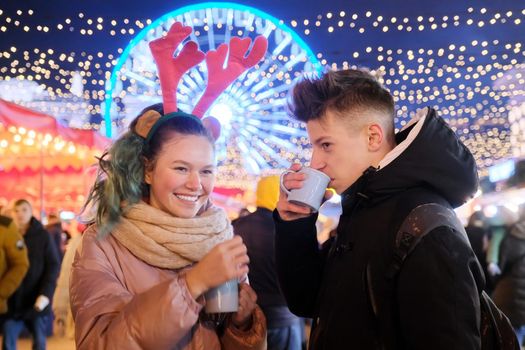 Christmas time, New Year holidays. Young people, couple of teenagers having fun at Christmas market, drinking hot tea from mugs, talking, laughing, background garlands of evening city, Ferris wheel