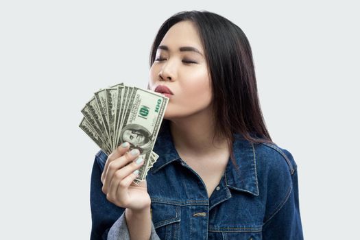 Loving money. Portrait of satisfied attractive young businesswoman in blue denim jacket standing, kissing with closed eyes and demonstrating fan of money. Indoor,studio shot, isolated, gray background