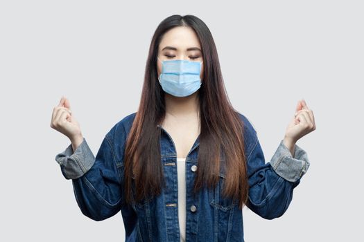 Portrait of calm beautiful brunette asian young woman with surgical medical mask in blue denim jacket standing with raised arms and yoga meditating. indoor studio shot, isolated on grey background.